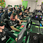 Dozens of Armada players sit in gaming chairs at desktop computers. They are wearing headphones and playing video games.
